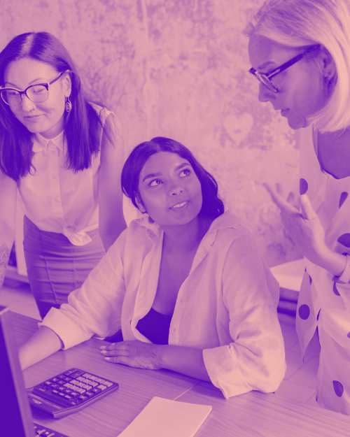 Three woman talking at work, Two are standing, one is sitting with a laptop in front of her.